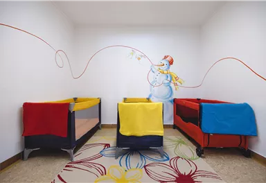 Ein freundlicher Raum mit drei Babybetten in bunten Farben. An der Wand ist ein fröhlicher Schneemann mit einer bunten Schnur abgebildet.