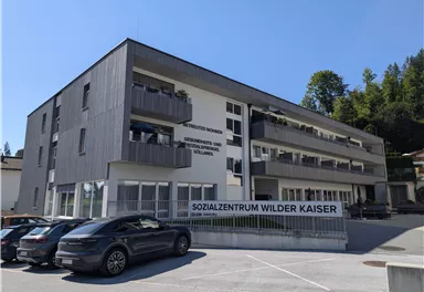 Ein modernes Sozialzentrum mit mehreren Etagen und Balkonen. Vor dem Gebäude stehen mehrere Autos und im Hintergrund sind Bäume zu sehen.