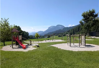 Ein schöner Spielplatz mit Rutsche und Schaukeln in einer grünen Landschaft. Im Hintergrund sind Berge und ein klarer Himmel zu sehen.