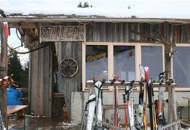 Eine rustikale Skihütte mit dem Schild „STALL-BAR“. Im Vordergrund stehen Skier auf einem Holzständer.