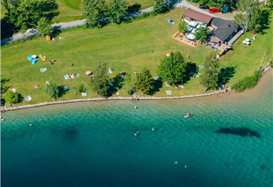 A beautiful lake with green grass and trees. People are enjoying the sun by the shore and in the water.