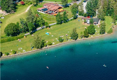 A picturesque lake with green grass and trees along the shore. In the background, there are some buildings and people relaxing by the water.