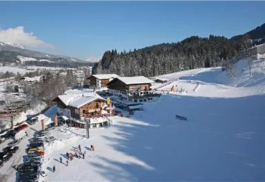 Eine malerische Berglandschaft mit schneebedeckten Hügeln und traditionellen Hütten. Im Vordergrund sind Skifahrer und Autos auf einem Parkplatz zu sehen.