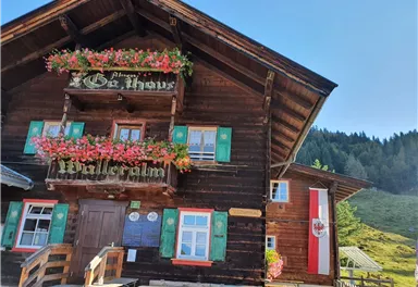 A traditional wooden house with flower-filled balconies in a picturesque mountain landscape. The Austrian flag is fluttering next to the entrance door.