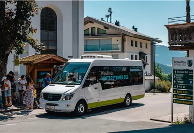 Ein kleiner Bus fährt durch eine sonnige Straße in einem Dorf. Im Hintergrund sind Häuser und einige wartende Personen zu sehen.