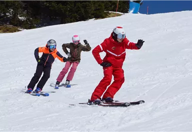 A group of skiers is going down a slope. The ski instructor in red clothing is leading the children on the slope.