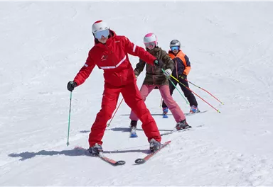 A ski instructor in red clothing shows students how to ski in the snow. The students are wearing ski helmets and are ready to learn the descent.