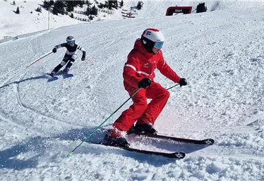 Two skiers are riding on a snow-covered slope. The skier in red clothing is carving a turn, while the one in blue and black is following behind him.