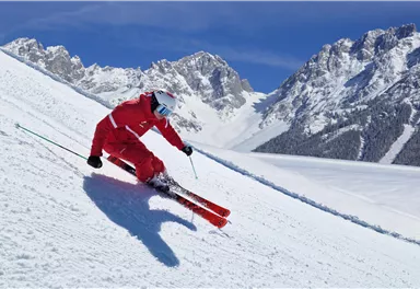 A skier in red is skiing down a snow-covered slope. In the background, majestic mountains and a clear blue sky can be seen.