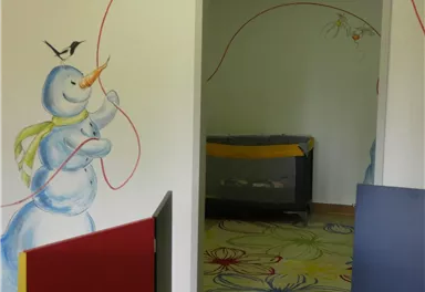 Ein freundlicher Schneemann mit einem roten Faden steht an einer Wand, während ein Fuchs darüber sitzt. Das Bild zeigt einen einladenden Raum mit bunten Möbeln und einem fröhlichen Bodenmuster.