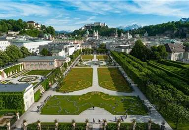 A beautiful park with artistically designed flower beds and lovely paths. In the background, historical buildings and mountains can be seen.