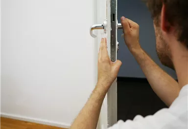 A man is installing a door handle on a door. The room has a bright floor and a neutral wall color.