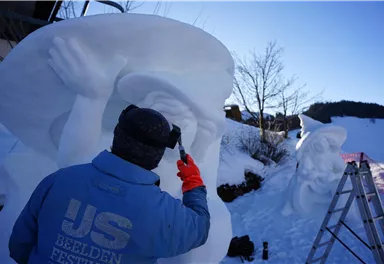Ein Künstler arbeitet an einer Schneeskulptur im Freien. Der Himmel ist klar und strahlend blau.