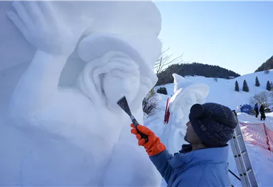 Ein Künstler schnitzt eine Schneeskulptur im Freien. Im Hintergrund sind weitere Schneeskulpturen und schneebedeckte Hügel zu sehen.