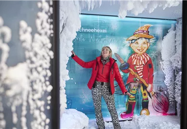 Eine Frau steht in einer winterlichen Szene mit Schnee und Farne. Im Hintergrund ist eine bunte Figur eines Schneemanns zu sehen.