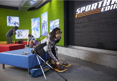 Eine Sportinfrastruktur mit Sitzmöbeln und einem Bildschirm. Eine Person bereitet sich darauf vor, Ski zu fahren.