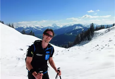 A man is hiking on a snow-covered mountain with ski poles. In the background, impressive mountains and a clear sky can be seen.