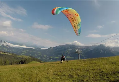 A person is paragliding over a green meadow and impressive mountain scenery. The sky is blue with a few clouds.