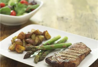 Ein Teller mit gegrilltem Steak, grünen Bohnen und gebratenen Kartoffeln. Im Hintergrund befindet sich eine Schüssel mit frischem Salat.
