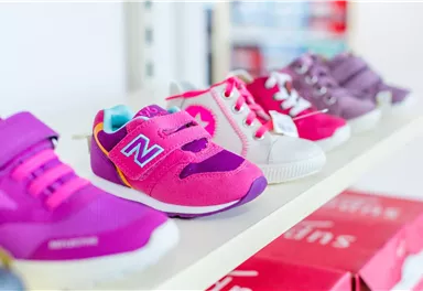 Colorful children's shoes in various designs are displayed on a shelf. The shoes are in the colors pink, purple, and white.