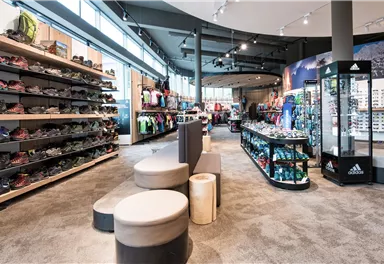 Ein gut sortierter Sportartikelgeschäft mit Regalen voller Schuhe und Bekleidung. Die Beleuchtung ist hell und der Raum wirkt modern und einladend.