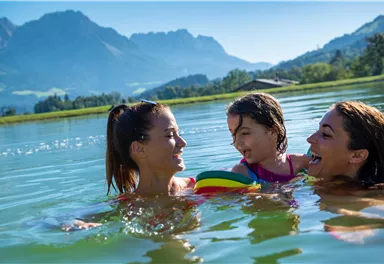 A sunny day by the water with two women and a child. They are having fun playing in the water with a colorful ball and a beautiful mountain landscape in the background.