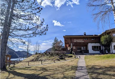 A picturesque inn by the lake, surrounded by trees and meadows. The clear sky and gentle landscape invite you to linger.