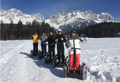 Eine Gruppe von fünf Personen fährt auf Schnee-Scootern in einer winterlichen Landschaft. Im Hintergrund sind beeindruckende Berge und ein klarer blauer Himmel zu sehen.