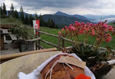 A freshly baked bread on a table, surrounded by colorful flowers. In the background, gentle hills and a cloudy sky can be seen.