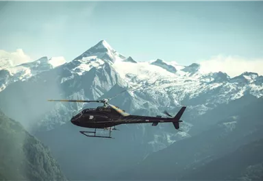 A helicopter flies over majestic mountains with snow-capped peaks. The sky is clear and the landscape is breathtaking.