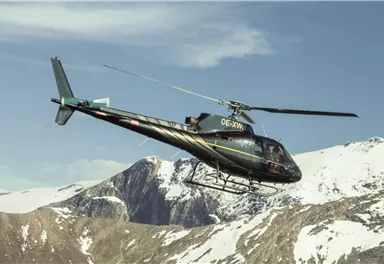A helicopter flies over snow-covered mountains. The sky is clear and blue.
