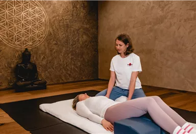 A woman is lying relaxed on a mat while another woman sits opposite her, taking care of her. In the background, a Buddha statue and a geometric wall design are visible.