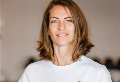 A woman with brown hair is smiling in a bright room. She is wearing a white T-shirt with the print "SHIATSU".
