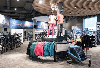 Ein Fahrradgeschäft mit verschiedenen Fahrrädern und Outdoor-Bekleidung. Zwei Schaufensterpuppen präsentieren Sportbekleidung auf einem zentralen Display.