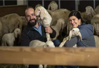 A man and a woman are each holding a lamb in their hands, surrounded by more sheep in a stable. Both are smiling kindly at the camera.