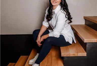 Eine lächelnde Frau sitzt auf einer Treppe aus Holz und Metall. Sie trägt ein weißes Hemd und blickt in die Kamera.