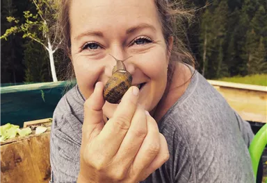 A woman is holding a small snail in her hand and smiling at the camera. In the background, green trees and a garden are visible.