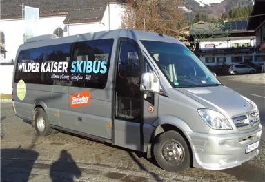 A silver bus with the inscription "Wilder Kaiser Skibus" is parked on a paved road. In the background, there are mountains and alpine architecture visible.