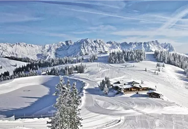 A picturesque winter landscape with snow-covered hills and mountains. In the center stands an alpine chalet surrounded by snow-covered trees.
