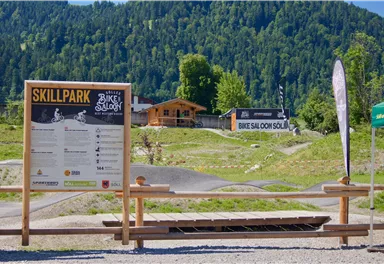 A skill park for mountain bikers with an information sign. In the background, you can see the surrounding mountains and some buildings.