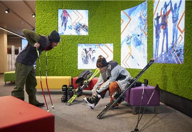 Zwei Personen in einer modernen Umgebung bereiten sich zum Skifahren vor. Hinter ihnen sind farbige Sitzmöbel und Bilder von Bergaktivitäten zu sehen.