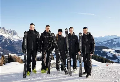 A group of five people is standing in the snow with skis in their hands. In the background, mountains and a clear sky can be seen.