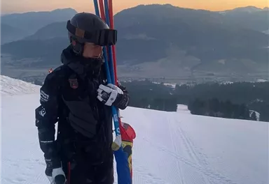 Ein Skifahrer steht auf einer verschneiten Piste mit Skistöcken in der Hand. Im Hintergrund sind die Berge und der Sonnenuntergang zu sehen.