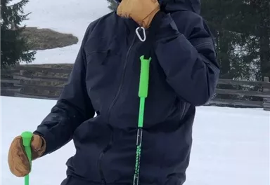 Ein fröhlicher Skifahrer mit Helm und Handschuhen steht auf der Piste. Im Hintergrund sind verschneite Bäume und neblige Berge sichtbar.