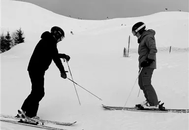Zwei Skifahrer stehen auf einer schneebedeckten Piste. Der Himmel ist grau und die Umgebung wirkt winterlich.