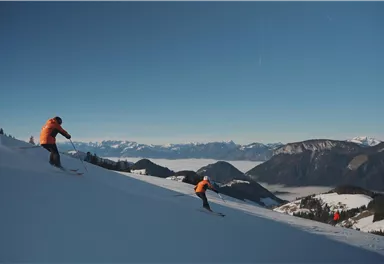 Zwei Skifahrer fahren eine breite Piste hinunter. Im Hintergrund sind schneebedeckte Berge und ein klarer blauer Himmel zu sehen.