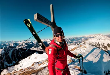 A skier stands on a summit overlooking snow-covered mountains. The sky is clear and blue.