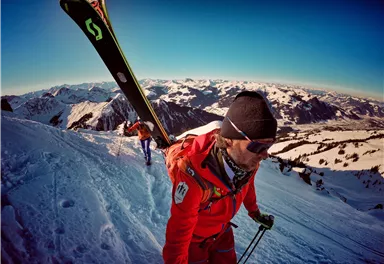 A person in a red ski suit is hiking with skis on a snow-covered mountain. In the background, stunning alpine landscapes can be seen.