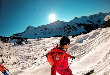 A skier in red is skiing through a snowy landscape. In the background, majestic mountains and a clear sky can be seen.