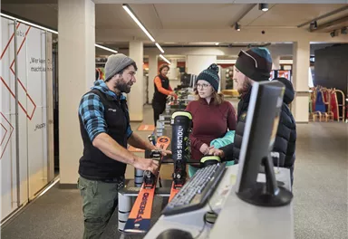 Ein Skiverleih mit Mitarbeitern, die Ausrüstungen an Kunden erklären. Zu sehen sind Skis und Winterbekleidung in einem modernen Umfeld.